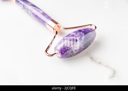 close up of facial massage amethyst gua sha face roller on white marble background. Face roller, massager made from natural stones, home spa. Stock Photo