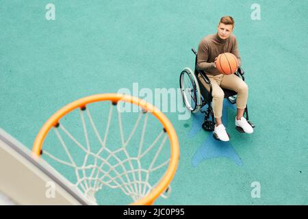 Serious young handicapped man sitting in wheelchair and playing basketball on sports ground Stock Photo