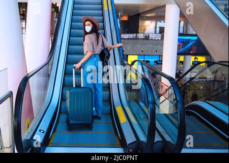 Young beautiful woman wearing mask uses moving stairway at the airport. Travel concept Stock Photo