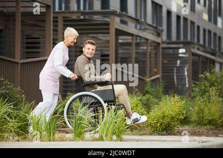 Positive energetic lady in medical uniform pushing wheelchair with boy while they walking in city Stock Photo