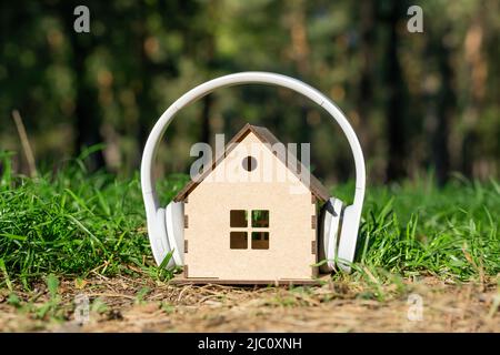 White wireless headphones on a miniature wooden house model in the woods. Natural house noise and acoustic insulation concept. Stock Photo