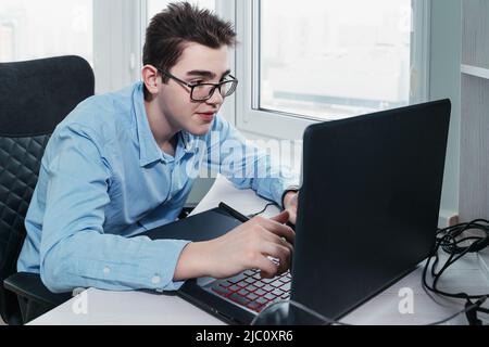 young man in glasses as he works on his laptop using a graphics tablet Stock Photo