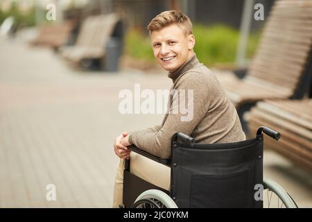 Portrait of smiling handsome young handicapped man in brown sweater sitting in wheelchair outdoors Stock Photo