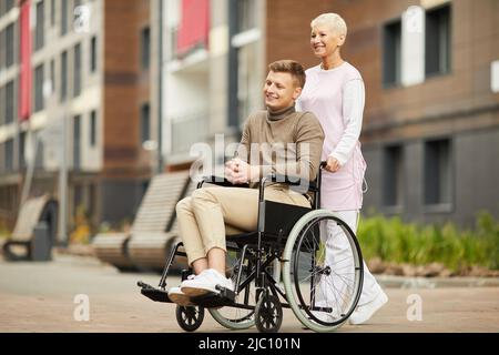 Positive handsome young man in casual outfit undergoing rehabilitation in medical center, nurse pushing wheelchair with young man Stock Photo