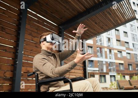 Content satisfied young wheelchair man in casual clothing gesturing hands and watching environment in VR headset while using simulator Stock Photo