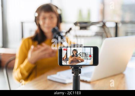 Female vlogger live streaming podcast using microphone, laptop and cellphone on tripod. Selective focus on smartphone camera screen with woman podcaster recording and broadcasting live video. Close-up Stock Photo