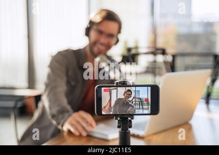 Male trendy podcast creator recording video podcast on smartphone. Selective focus on smartphone camera screen with man podcaster vlogger in headphones and glasses shooting live video for his channel Stock Photo