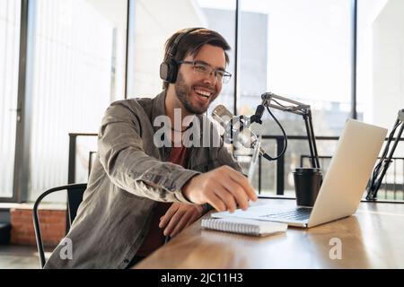 Young man host in headphones and glasses enjoying podcasting in studio, speaking into a microphone, holding a pen, using laptop. Handsome podcaster laughing while streaming live audio podcast Stock Photo