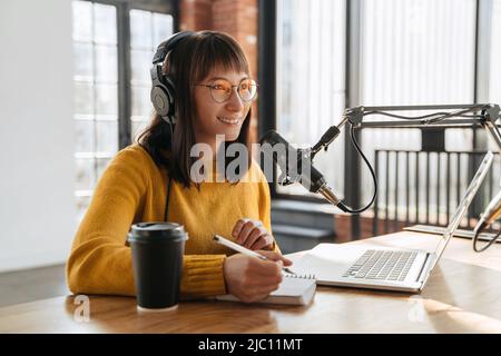 Podcasting and blogging. Young smiling woman broadcasting an audio podcast for her online show in studio, looking toward, using microphone, laptop and headphones, holding a pen, writing in notepad Stock Photo