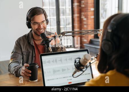 Female podcast creator interviewing a handsome smiling male guest while recording audio podcast in studio. Man and woman podcasters enjoying podcasting, talking to each other, speaking in microphone Stock Photo