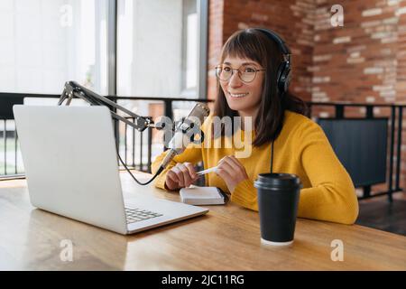 Young smiling woman in eyeglasses recording and broadcasting her podcast in studio. Female radio host in headphones recording live audio podcast using microphone and laptop, holding a pen in her hands Stock Photo