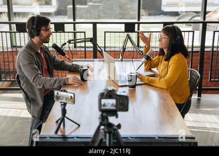 Two young bloggers, man and woman in headphones and glasses looking at each other while talking, recording conversation, interview for video blog. Female host interviewing guest for her video podcast Stock Photo