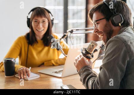 Selective focus of cheerful radio host laughing while recording podcast with colleague. Two radio hosts laughing while sitting near microphones in broadcasting studio and making audio podcast Stock Photo