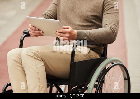 Close-up of unrecognizable disabled man in brown outfit sitting in wheelchair and browsing internet on tablet outdoors Stock Photo