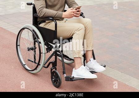Unrecognizable handicapped man sitting in wheelchair and texting sms on smartphone while walking in city Stock Photo