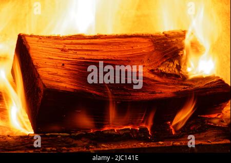 Flames lick around the base of a block of wood on an open fire, flames are roaring behind it. Stock Photo