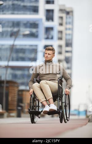 Serious pensive young man in casual sweater turning handrails while using wheelchair outside Stock Photo