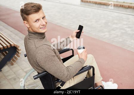 Portrait of cheerful handsome disabled man in brown turtleneck sitting in wheelchair outside and using smartphone while drinking coffee Stock Photo