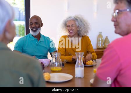 Happy multiracial senior friends talking while having breakfast at dining table in retirement home Stock Photo