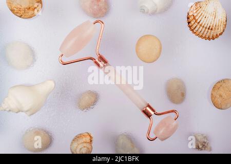 Rose quartz face roller and sea shells with water drops on white background. Selfcare concept. Top view, flat lay. Stock Photo