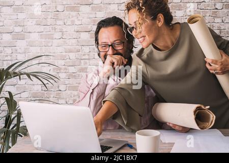 Happy couple of adult worker smile and enjoy job together. Woman pointing at computer display and man laugh. Adult people with laptop and papers. Coll Stock Photo