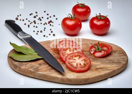 Slices of ripe tomatoes on a cutting board with a knife and three whole tomatoes nearby, bay leaf and pepper on a pale blue background. Eating at home Stock Photo