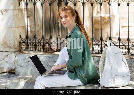 redhead freelancer sitting on concrete bench and using laptop near backpack Stock Photo
