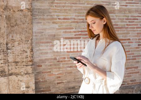 young redhead woman messaging on smartphone near brick wall on urban street Stock Photo