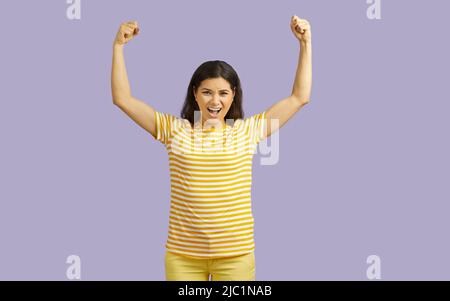 Portrait of funny proud young woman raised her hands up posing like athlete, demonstrating her power, isolated over purple background Stock Photo