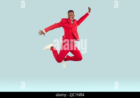 Happy funny young african american man in red suit high jumps on pastel light gray background. Stock Photo