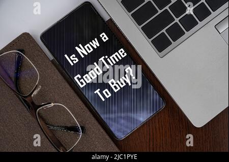 Is now a good time to buy? Top view of phone on table near laptop, notepad and glasses with inscription. Stock Photo