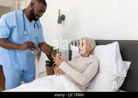 African american male health worker giving medical pills to caucasian senior woman lying on the bed Stock Photo