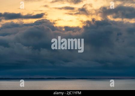Dramatic storm clouds above sea water surface with golden sunrise behind the clouds on a warm day of early autumn. Stock Photo