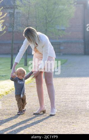 Portrait of cute small caucasian baby boy learn walking at the park holding mom hands, little toddler infant child make do first steps with mother support, childcare, upbringing concept. High quality photo Stock Photo