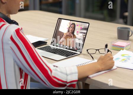 Asian businesswoman on video call with asian female colleague writing on diary at desk in office Stock Photo