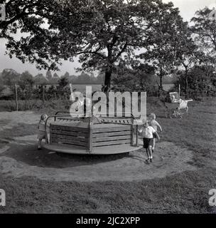 1950s, historical, outside in a countryside playground, young children playing on a wooden roundabout, England, UK. A coach-built pram of the era is sat on the grass. Stock Photo