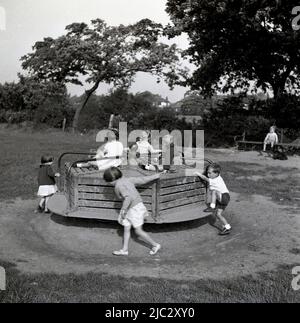 1950s, historical, outside in a countryside playground, young children playing on a wooden roundabout, England, UK. Stock Photo