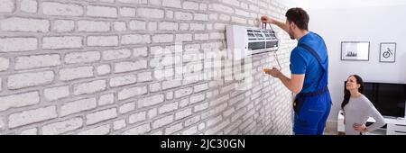 Smiling Woman Looking At Male Technician Repairing Air Conditioner With Digital Multimeter At Home Stock Photo
