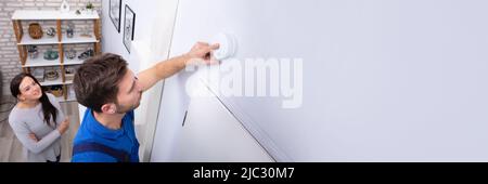 Pretty Woman Standing Near Repairman Installing Smoke Detector On Wall At Home Stock Photo