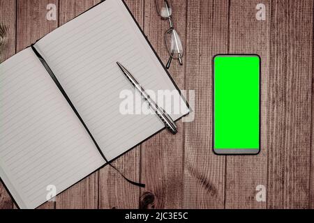 mobile phone with a green screen, a notepad and a pen on the background of wooden boards. Stock Photo