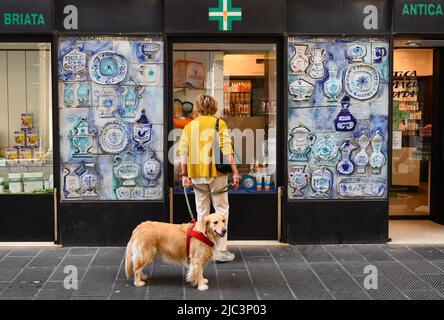 A lady with a dog on a leash outside a historic pharmacy decorated with ceramic tiles in the neighborhood of San Vincenzo, Genoa, Liguria, Italy Stock Photo