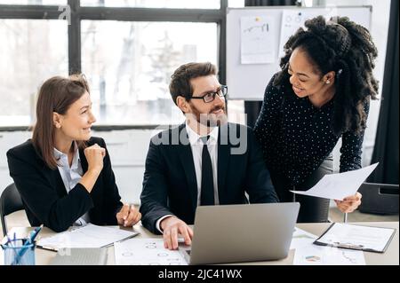 Multiracial colleagues are brainstorming on strategy together, sitting in modern office, studying documents, using laptop, discussing business plan, african american girl explaining information Stock Photo