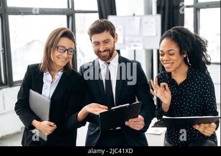 A team of business creative successful multiracial people, business partners, are standing in their modern office, happily discussing the project, predicting its success, discussing new ideas, smiling Stock Photo