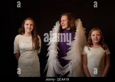 Mother with curly hair and cute teenagers girls with white wings looking like a nice angels posing in photo shoot in studio with black background Stock Photo