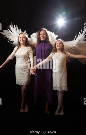 Mother with curly hair and cute teenagers girls with white wings looking like a nice angels posing in photo shoot in studio with black background Stock Photo