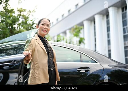 Elegant aged asian businesswoman in formal suit getting out of the car with happy facial expression. Stock Photo