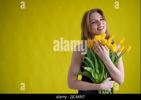 Caucasian woman with an armful of yellow tulips on a yellow background. International Women's Day. Bouquet of spring flowers Stock Photo