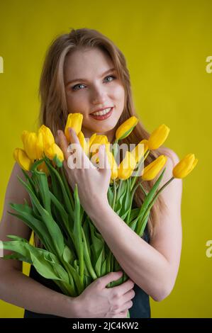 Caucasian woman with an armful of yellow tulips on a yellow background. International Women's Day. Bouquet of spring flowers Stock Photo
