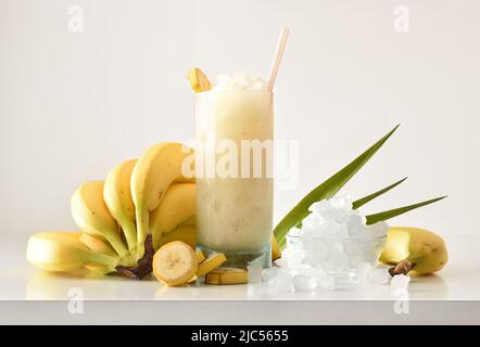Banana slush drink in tall glass with fruit and crushed ice around it on white table with isolated background. Front view. Horizontal composition. Stock Photo