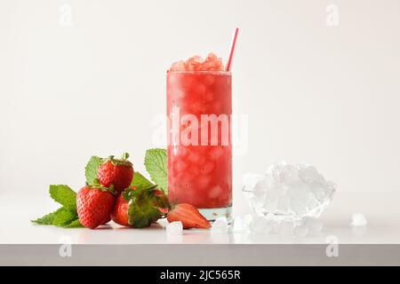 Strawberry slush drink in tall glass with fruit and crushed ice around it on white table with isolated background. Front view. Horizontal composition. Stock Photo